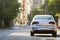 Close-up back view of new shiny expensive silver car moving along city street on blurred trees, cars and buildings background on