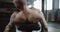 Close-up back view young bald athletic man lifting heavy dumbbells on bench, getting tired and resting in large gym.