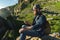 Close-up of a bearded man in jeans clothes in sunglasses and a cap with a backpack sitting at the foot of the epic rocks