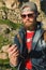 Close-up of a bearded man in jeans clothes in sunglasses and a cap with a backpack sitting at the foot of the epic rocks