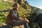 Close-up of a bearded man in jeans clothes in sunglasses and a cap with a backpack sitting at the foot of the epic rocks