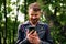 Close-up of a bearded man with red hair smiling while typing a message in a smartphone while standing amongst a forest