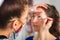 Close-up of a beautician wearing colorful protective mask during COVID-19 while applying make up on beautiful young woman who\'s si