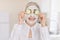 Close up of beautiful retired woman with facial mask on her face holding slices of fresh cucumber covering her eyes, on