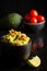 Close up of black bowl with guacamole on shiny black background with avocados, tomatoes and lime