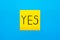 Close up black handwritten inscription yes word on one yellow square sticker on blue background with copy spase, horizontal