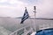 Close up of blue and white Greek flag on flagpole flying in wind. Torn Greek national flag waving on bow of ship against blue sea.