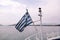 Close up of blue and white Greek flag on flagpole flying in wind. Torn Greek national flag waving on bow of ship against blue sea.