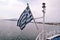 Close up of blue and white Greek flag on flagpole flying in wind. Torn Greek national flag waving on bow of ship against blue sea.