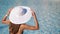 Close-up. Blurred water background. A young woman in big white hat sits near the pool and keeps her hat. With place for