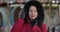 Close-up of brunette Caucasian woman in red coat and hood looking at camera and smiling. Positive lady standing on city