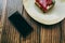 Close up of cake with raspberry sauce on plate, cup of cappuccino, mobile phone with blank empty screen lying on wooden