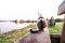 Close-up of a cat from behind sitting on a bench by a lake facing a blurred woman sitting in the background looking at him -