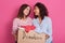 Close up of cheerful helpful volunteer girls standing isolated over pink background, holding box with different clothes. Full box