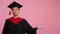 Close-up of a cheerful student in a hat and gown. Copy space on pink background
