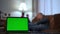 Close-up chromakey laptop with blurred Caucasian man sitting on couch at background reading book. Unrecognizable relaxed