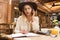 Close up of a confused girl in hat sitting at the cafe table indoors, talking on mobile phone, taking notes in a notebook