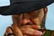 Close-up, a cowboy plays the harmonica in his mouth with his hat covering his eyes.