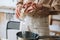 Close-up cropped shot of female artisan adding white dry soy wax in glass jar into pot of boiling water for creating