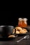 Close-up of cup of coffee, plate with cookies, spoon and jar of jam on dark table, black background, vertical,