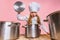 Close-up cute little girl in white cook uniform and huge chef& x27;s hat at kids kitchen with big pans isolated on pink