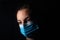 Close-up of a dark portrait of a very dimly lit woman. The woman is wearing a surgical mask, her gaze is lost on the horizon and