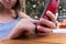 Close up detail of girl`s finger without nail polish touching screen of smartphone with red cover in table outdoors. Trees and