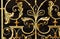 A close up of the detailed metal work of a gold wrought iron gate against a black background