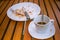 Close up dirty coffee cup and spoon setting on white saucer, knife and fork on white dirty dish. They are on wooden table.