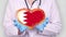 Close-up. Doctor in medical white coat, blue gloves holds in hands drawn pulsating heart with Bahrain flag. Concept of