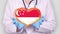 Close-up. Doctor in medical white coat, blue gloves holds in hands drawn pulsating heart with Singapore flag. Concept of