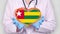 Close-up. Doctor in medical white coat, blue gloves holds in hands drawn pulsating heart with Togo flag. Concept of