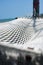 Close up of an empty white rope hammock hanging with trees in a shadow of tree branches on a tropical beach at noon.