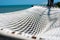 Close up of an empty white rope hammock hanging with trees in a shadow of tree branches on a tropical beach at noon.