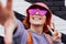 Close-up excited redhead woman in magenta heart-shaped sunglasses smiling and showing V finger sign while taking a