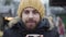 Close-up face of bearded Caucasian guy in yellow hat enjoying hot coffee outdoors. Young handsome man with brown eyes