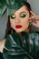 Close up face of beautiful young woman covering by green monstera green palm. Portrait of beauty woman with makeup red lips clear