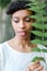 Close up face of black female person keeping green leaf, having bangs and wearing white blouse.