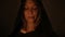 Close-up face of a girl in a black magic hood with eyes closed by candlelight in a dark room. Halloween Holidays and