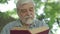 Close-up face of grey-haired bearded old man with brown eyes reading book in sunlight outdoors. Portrait of absorbed