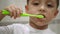 Close-up of the face of a little boy who holds a toothbrush and brushes his teeth. Mom`s hand puts white toothpaste on