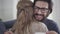 Close-up face of young caucasian man with black beard imitating joy as his wife showing affirmative pregnancy test, and