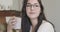 Close-up face of a young thoughtful Caucasian girl in elegant eyeglasses sitting with a cup of tea or coffe and looking