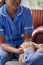 Close Up Of Female Care Worker In Uniform Holding Hands Of Senior Man Sitting In Care Home Lounge