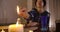 Close-up of female Caucasian hand lighting up candle standing at the foreground. Old serious fortune teller sitting at