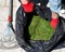 Close-up of female gardener wearing red and black gloves holding a black bag of mowed grass. Rake on the ground