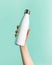 Close-up of female hand holding white reusable steel stainless thermo water bottle  on background of cyan, aqua menthe.