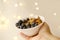 close-up female hand holds healthy food white cup, oatmeal, blueberries, nuts, almonds, peanuts, raisins, dried fruits on blurred