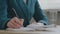 Close up female hands, caucasian woman in green jacket sitting at desk holding pen making notes in diary. Unrecognizable