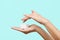 Close up of female hands holding moisturizer cream and dipping finger in it, isolated over pastel blue background.